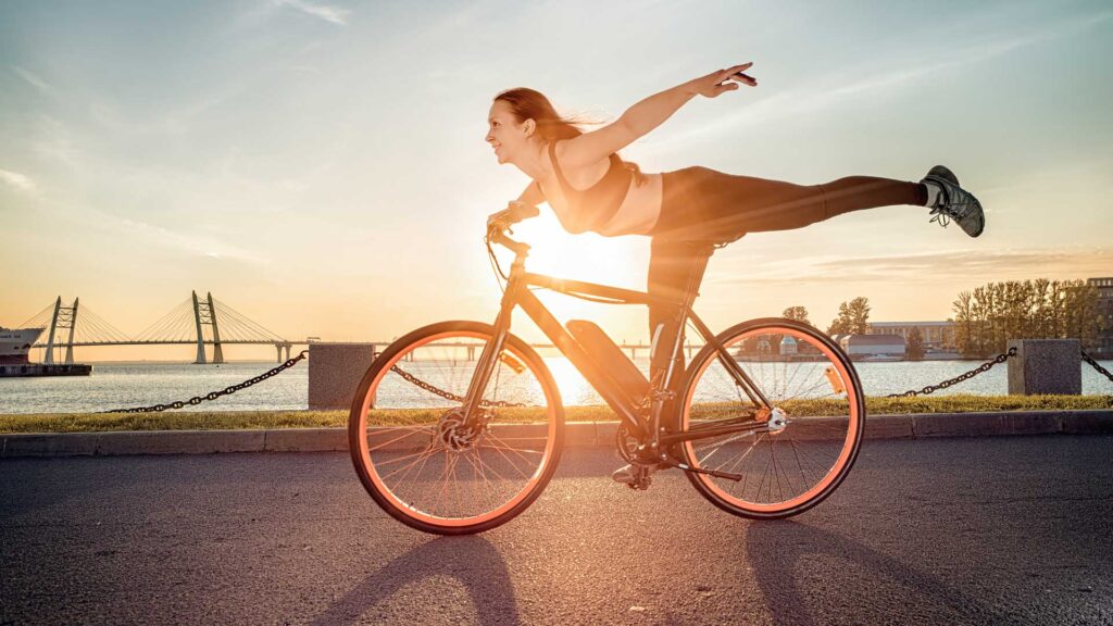 Woman in fitness clothing happily cycling, one leg stretched out and arms open - cycling is healthy and frees you up.