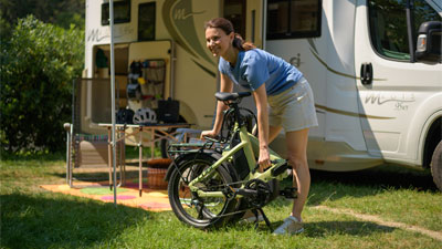 Zu sehen ist eine Frau, welche vor einem Wohnmobil steht. Vor ihr steht ein eingeklapptes grünes Faltrad.