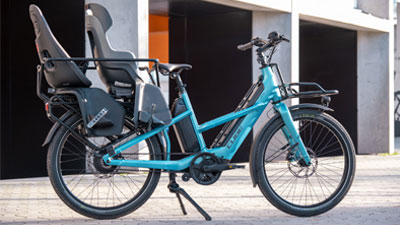 Zu sehen ist ein blaues Cube Longtail Hybride Lastenfahrrad, mit zwei ausgestatteten Kindersitzen. 