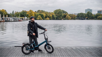 Ein Mann steht neben seinem blauen Faltrad auf einem Steg. Im Hintergrund ist Gewässer und eine Stadt zu sehen.